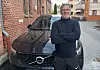 Mann i mørke klær står med korslagte armer foran en svart Volvo XC40 ved en mursteinsbygning.