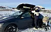 Person laster ski i skiboks på mørk Tesla parkert i snødekt fjellandskap ved barnevogn.